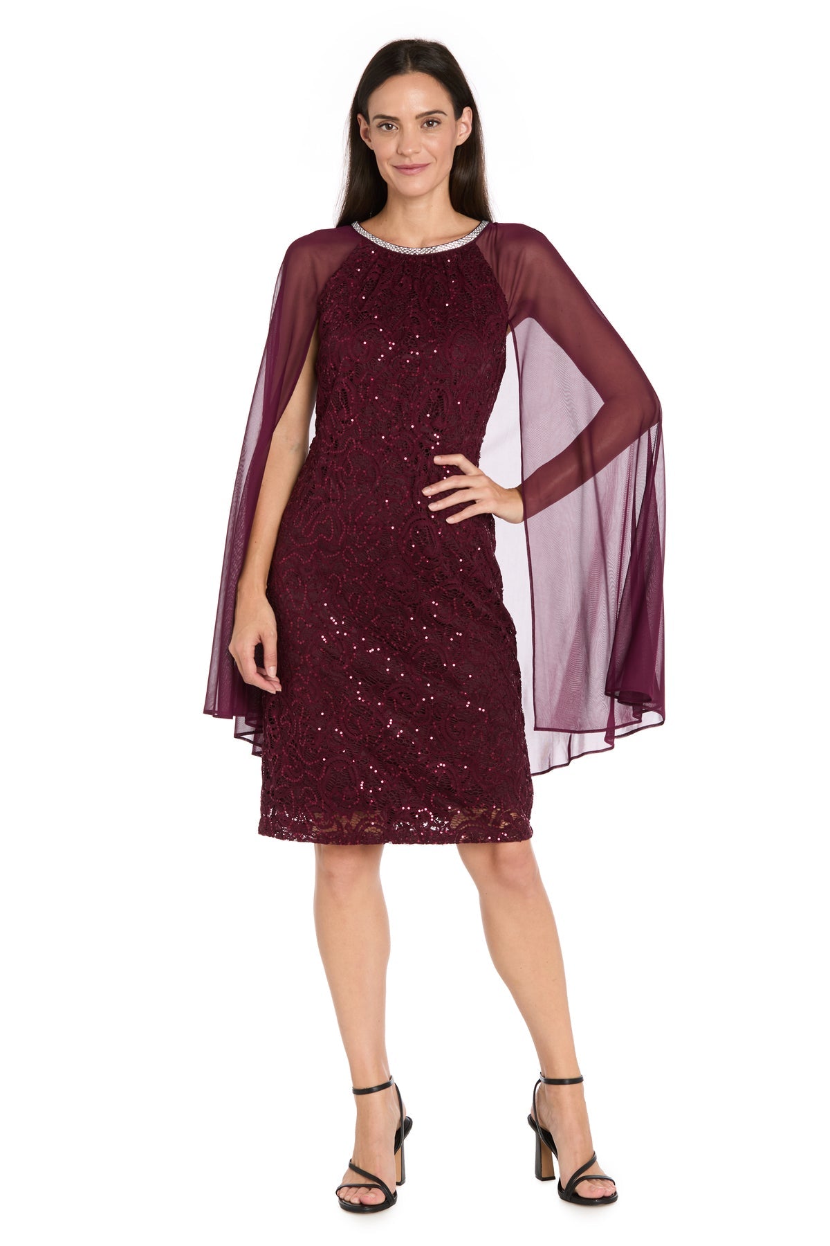 A woman stands against a white background wearing the Knee-Length Cape Party Dress: R&M Richards 3864, featuring burgundy sequin-lace, sheer cape sleeves, and black strappy heels.